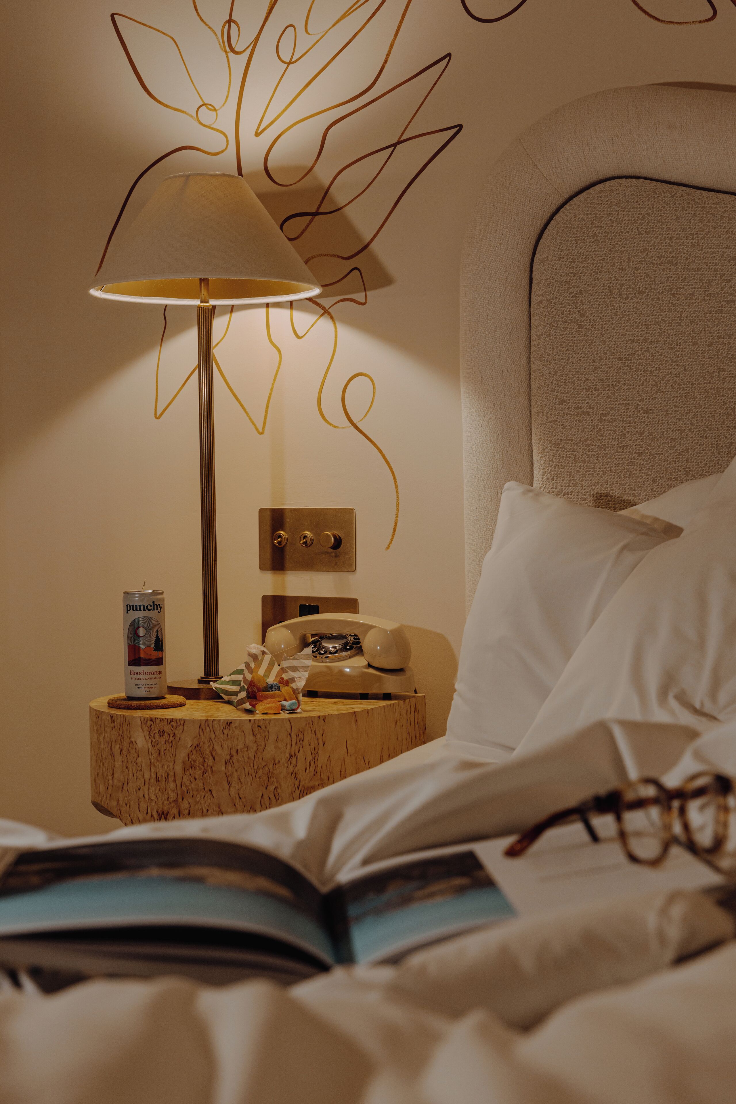 A neatly made bed with a book and glasses, a bedside lamp, a can of punch, and a telephone on a wooden nightstand.