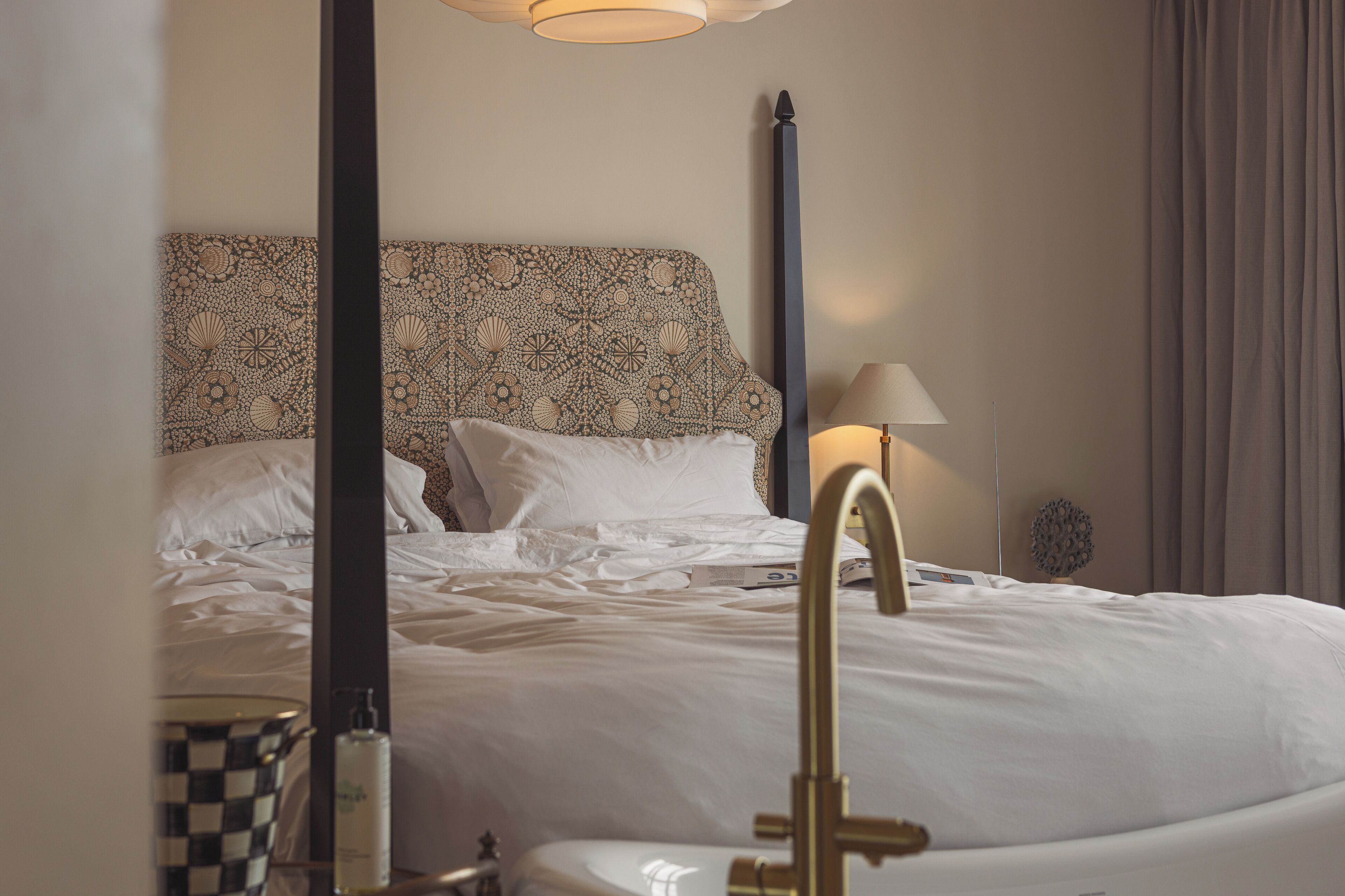 A neatly made bed with white linens, a patterned headboard, a bedside lamp, and a checkered nightstand with a bottle.