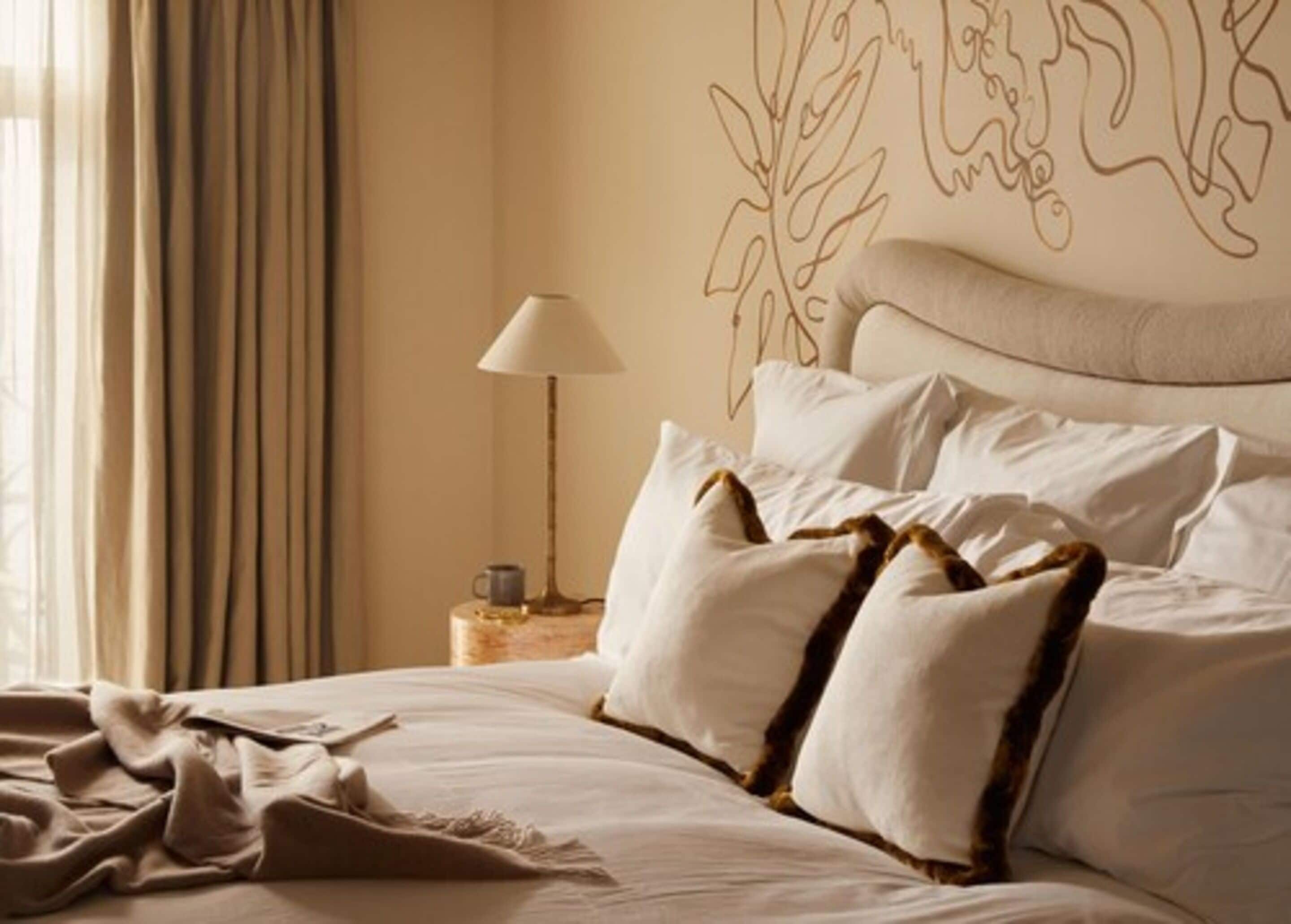 A neatly made bed with white linens and patterned pillows, a bedside lamp, and a wall with a simple line drawing.