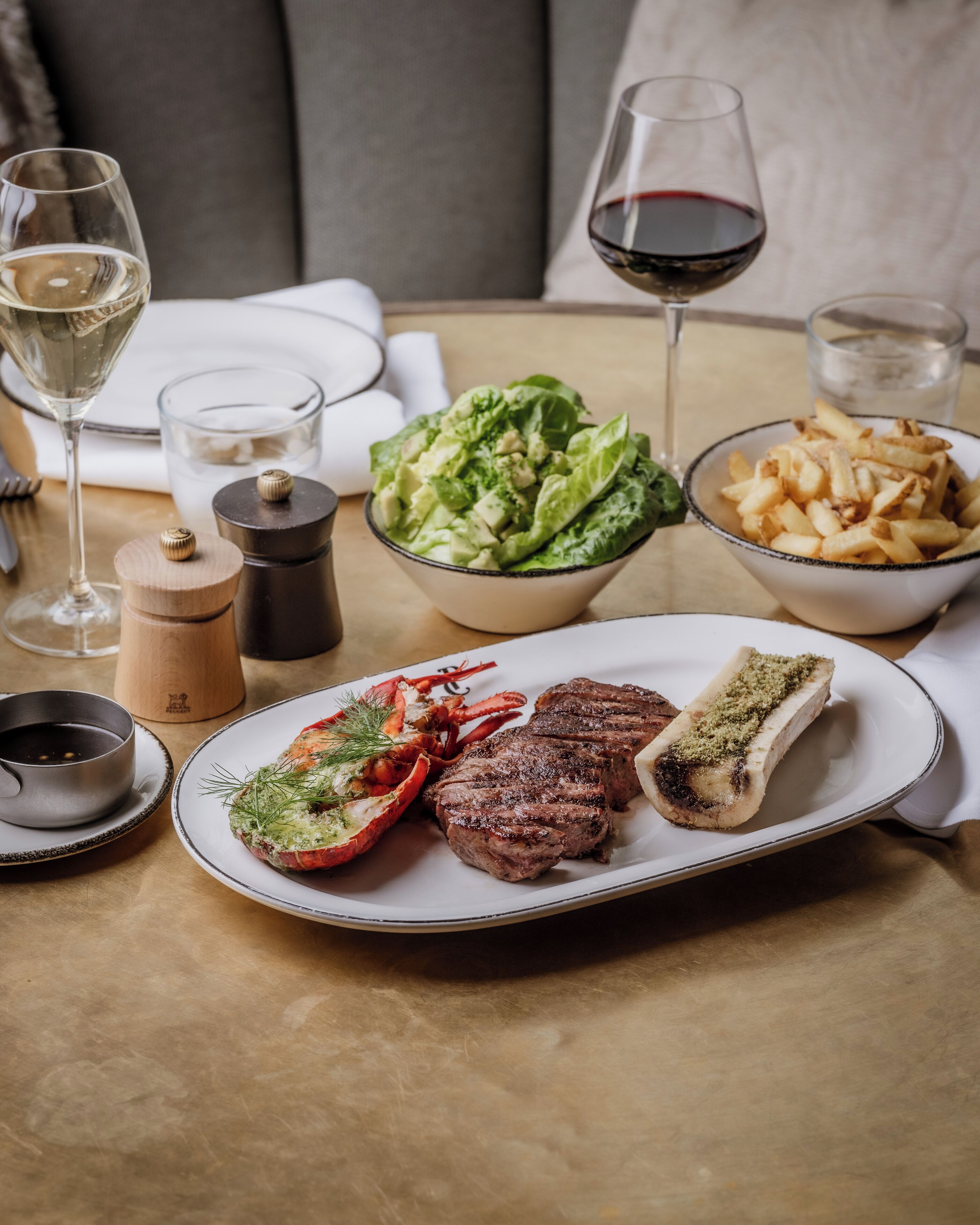 A dining table with a plate of steak, lobster, and vegetables, a bowl of salad, and a glass of red wine.