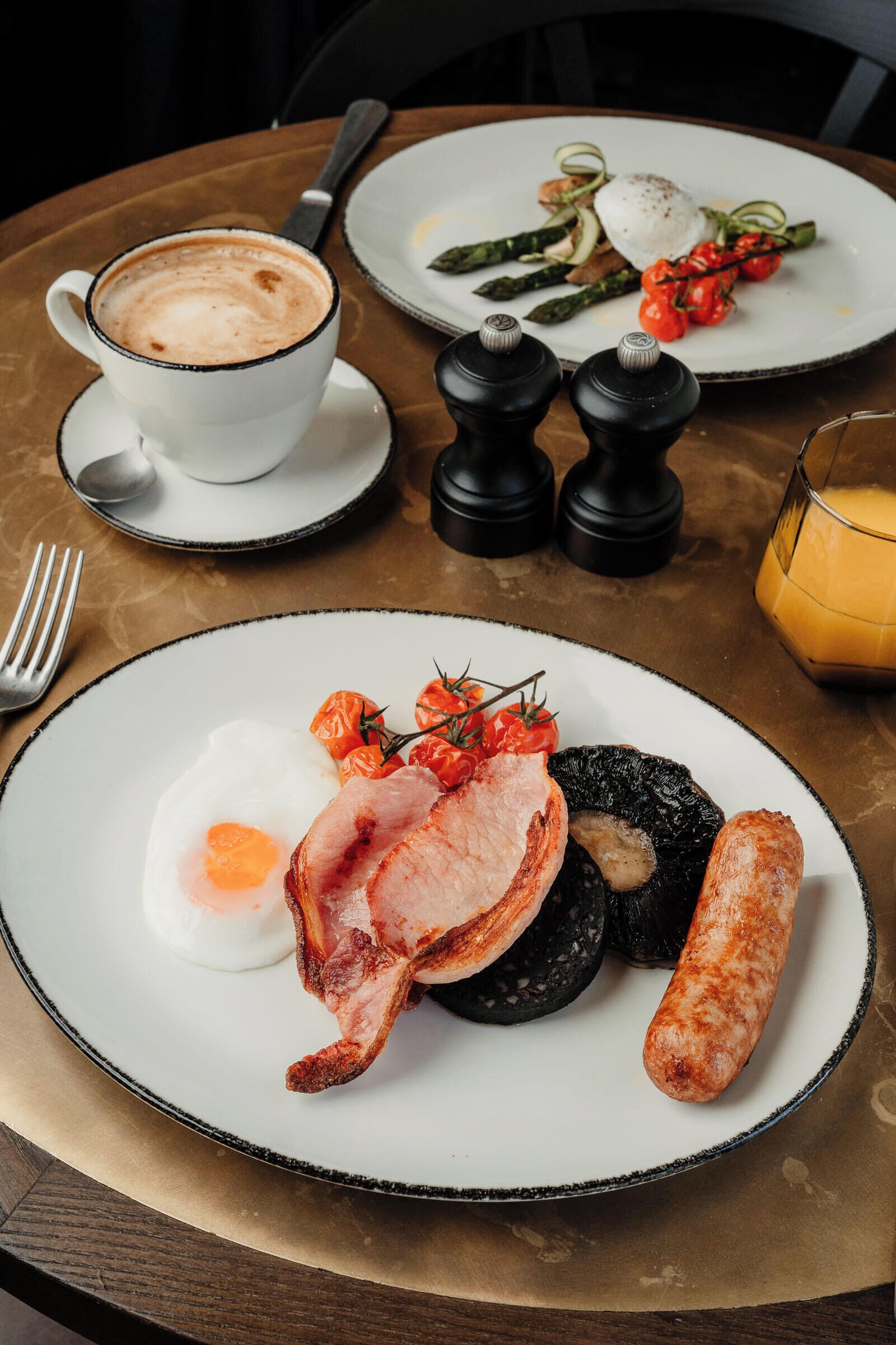 Un desayuno con platos de huevos, tocino, salchichas y verduras, una taza de café y un vaso de zumo de naranja.
