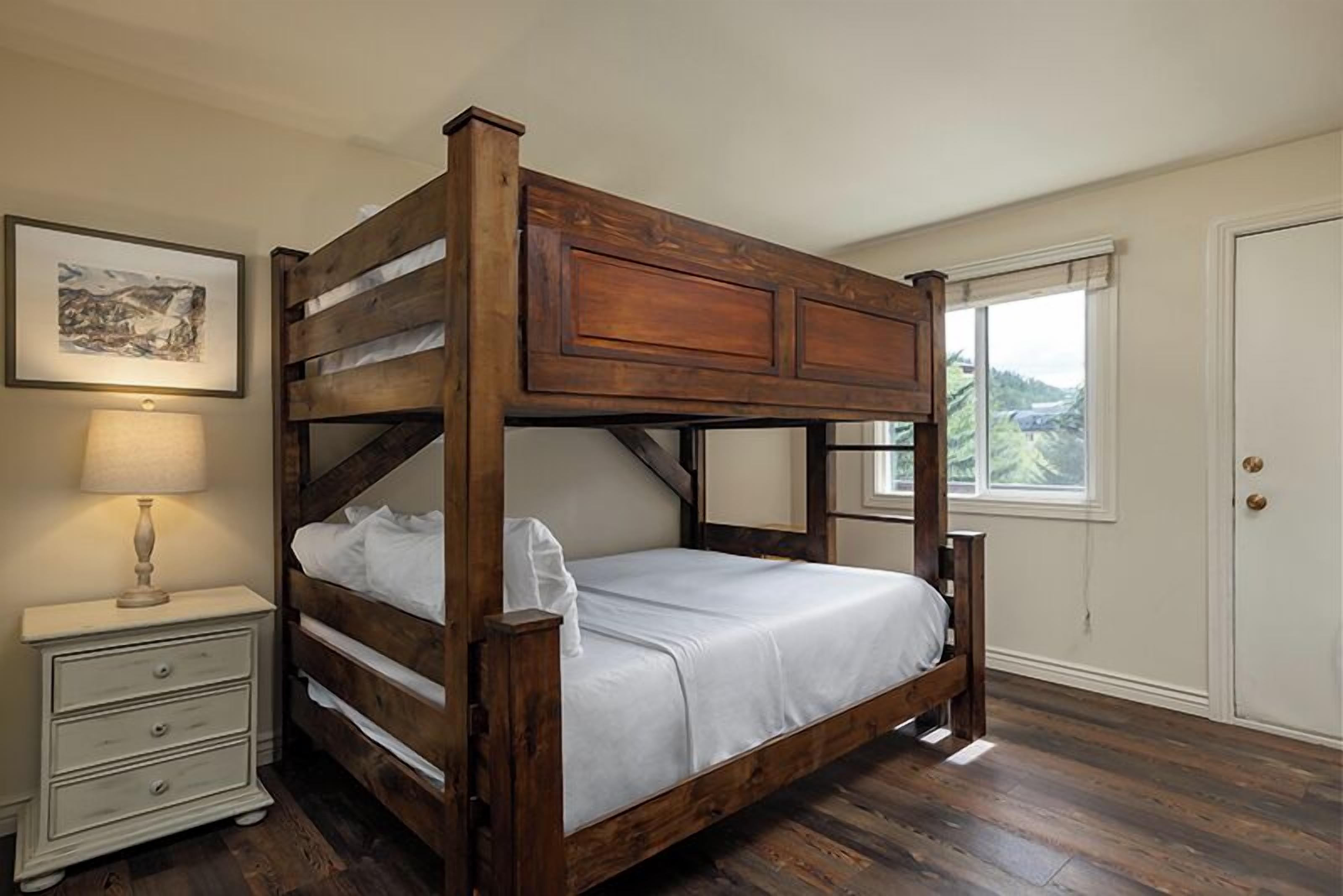 Ein hölzernes Himmelbett mit weißer Matratze, ein Nachttisch mit Schubladen, eine Lampe und ein Fenster mit Blick auf Grünflächen.