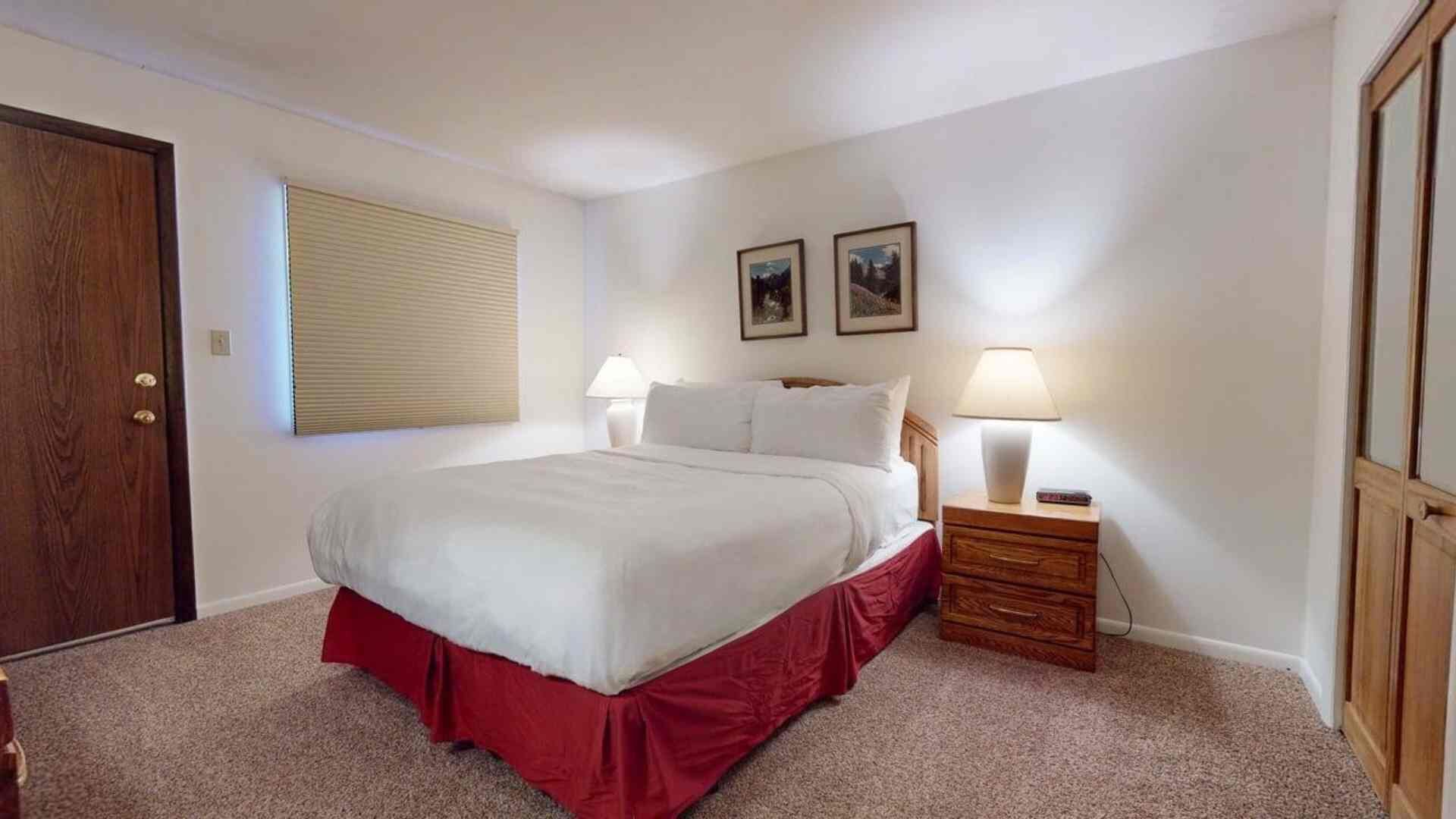 Ein Schlafzimmer mit einem Bett, einem Nachttisch, einer Lampe, einem Fenster mit Jalousien und zwei gerahmten Bildern an der Wand.