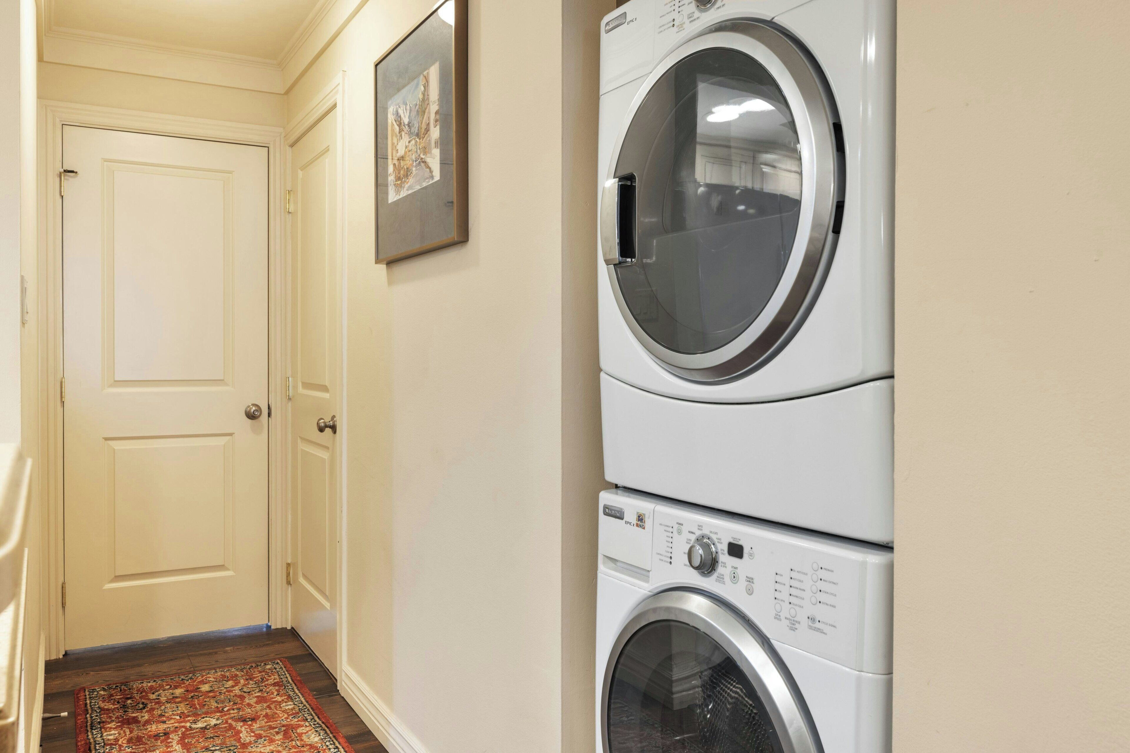 A laundry area with two stacked washing machines, a door, and a patterned rug.