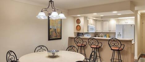 A dining area with a round table and six chairs, a chandelier, and a kitchen in the background.
