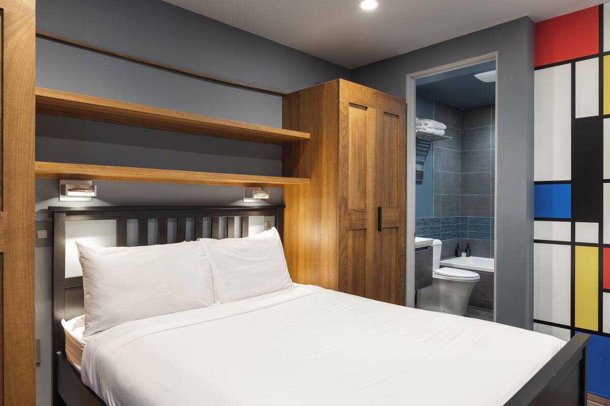 Une chambre à coucher avec un lit, une tête de lit en bois, des étagères intégrées et une vue sur une salle de bain équipée de toilettes et d’un lavabo.