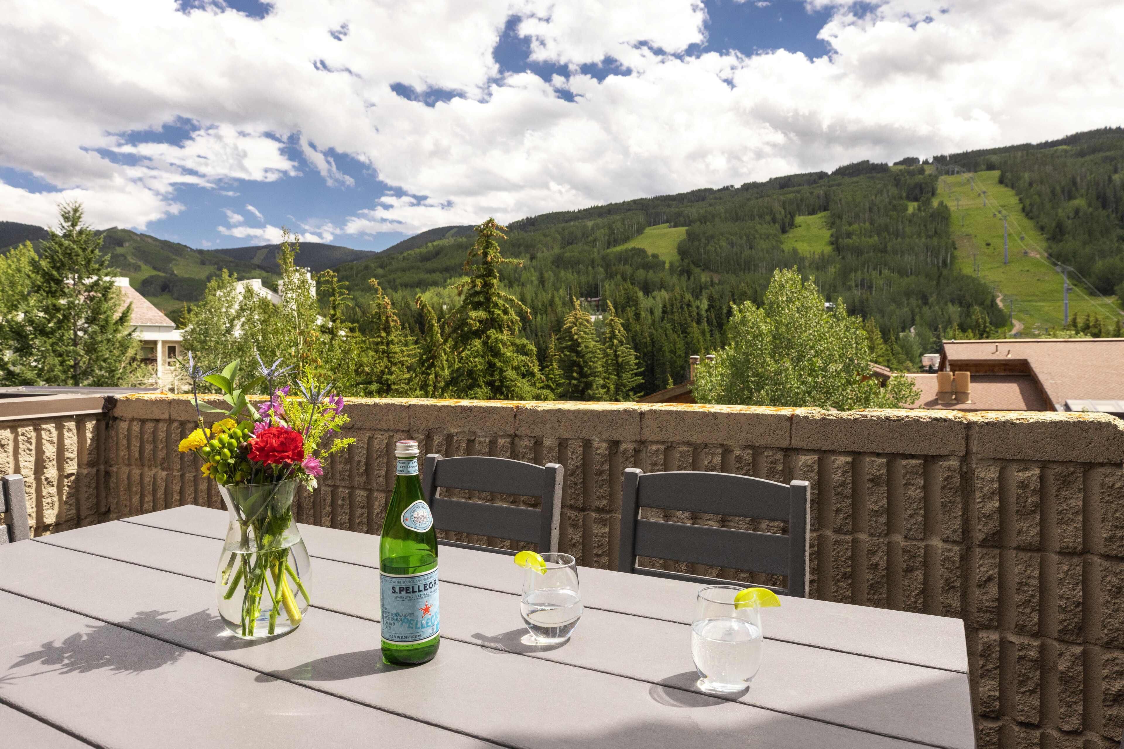 Uma mesa com uma garrafa de água S.Pellegrino, dois copos e um vaso de flores, com uma paisagem montanhosa ao fundo.