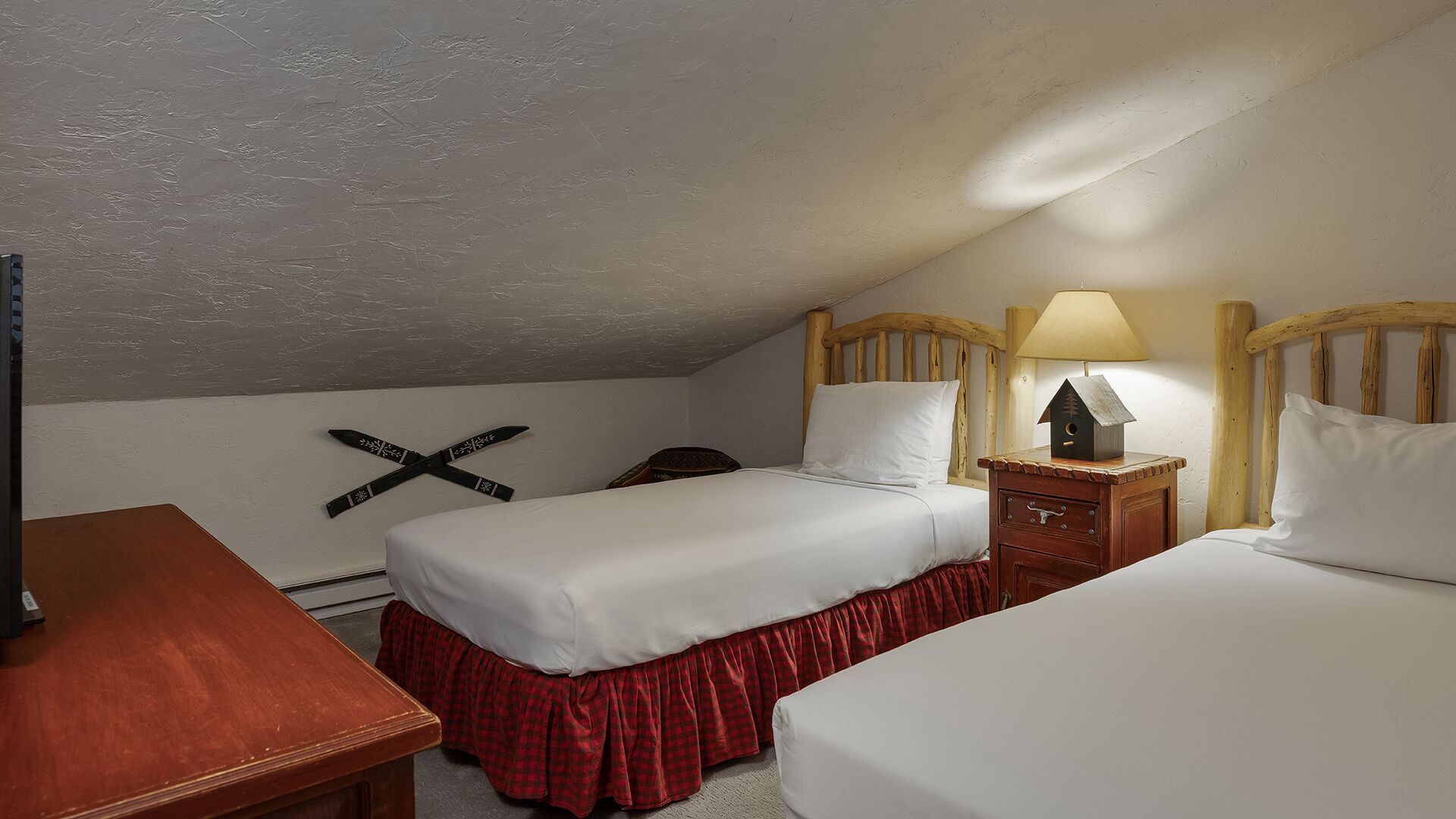 Une chambre avec deux lits simples, une table de chevet en bois avec une lampe, une tête de lit en bois et un crucifix fixé au mur.