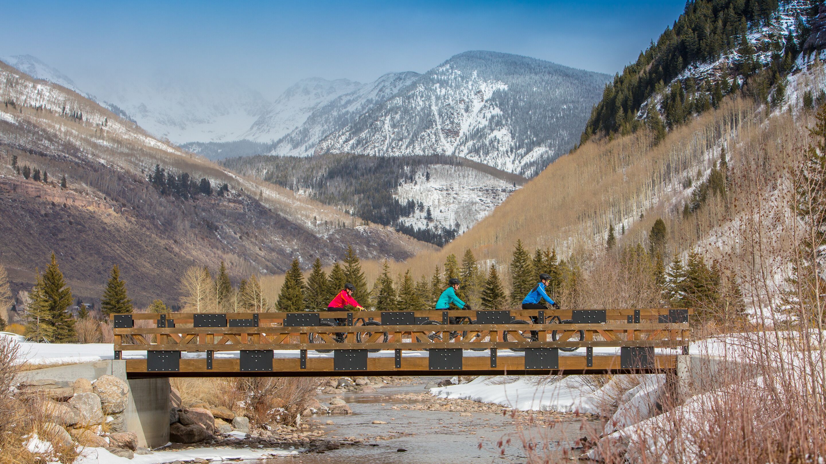 A wooden bridge over a river with snow-covered mountains in the background.
