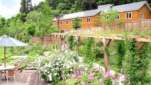 A wooden deck with a table and chairs, surrounded by lush greenery and flowering plants. - Pension Asanebou (Minakami)