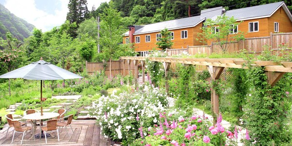 Una terraza de madera con mesa y sillas, rodeada de exuberante vegetación y plantas en flor.