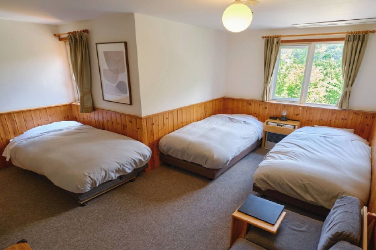 A room with three single beds, a small table, a chair, a framed picture, and a window with curtains.
