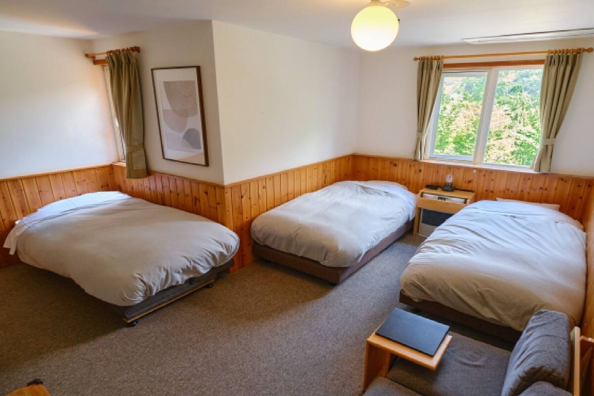A room with three beds, a small table, a chair, a window with curtains, and a framed picture on the wall.