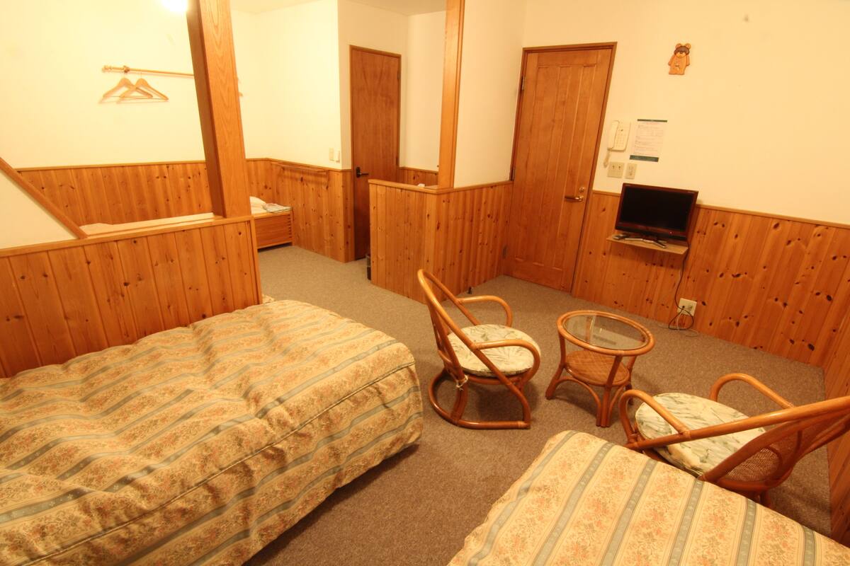 A room with two beds, a chair, a small table, a TV, and a wooden wall paneling.