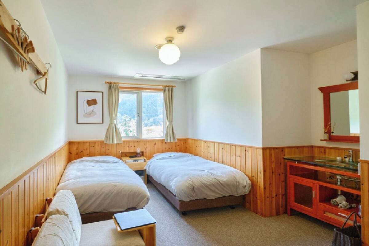 A room with two beds, a wooden bench, a small table, a mirror, and a window with curtains.