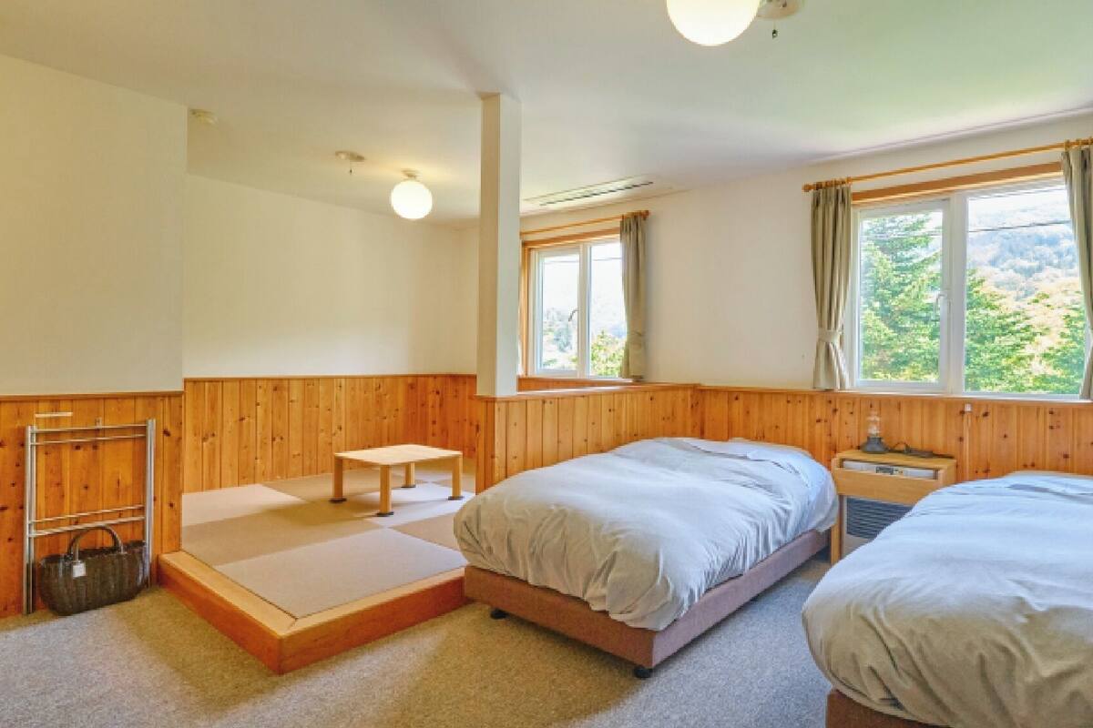 A room with two beds, a small table, a bench, and a window with curtains.