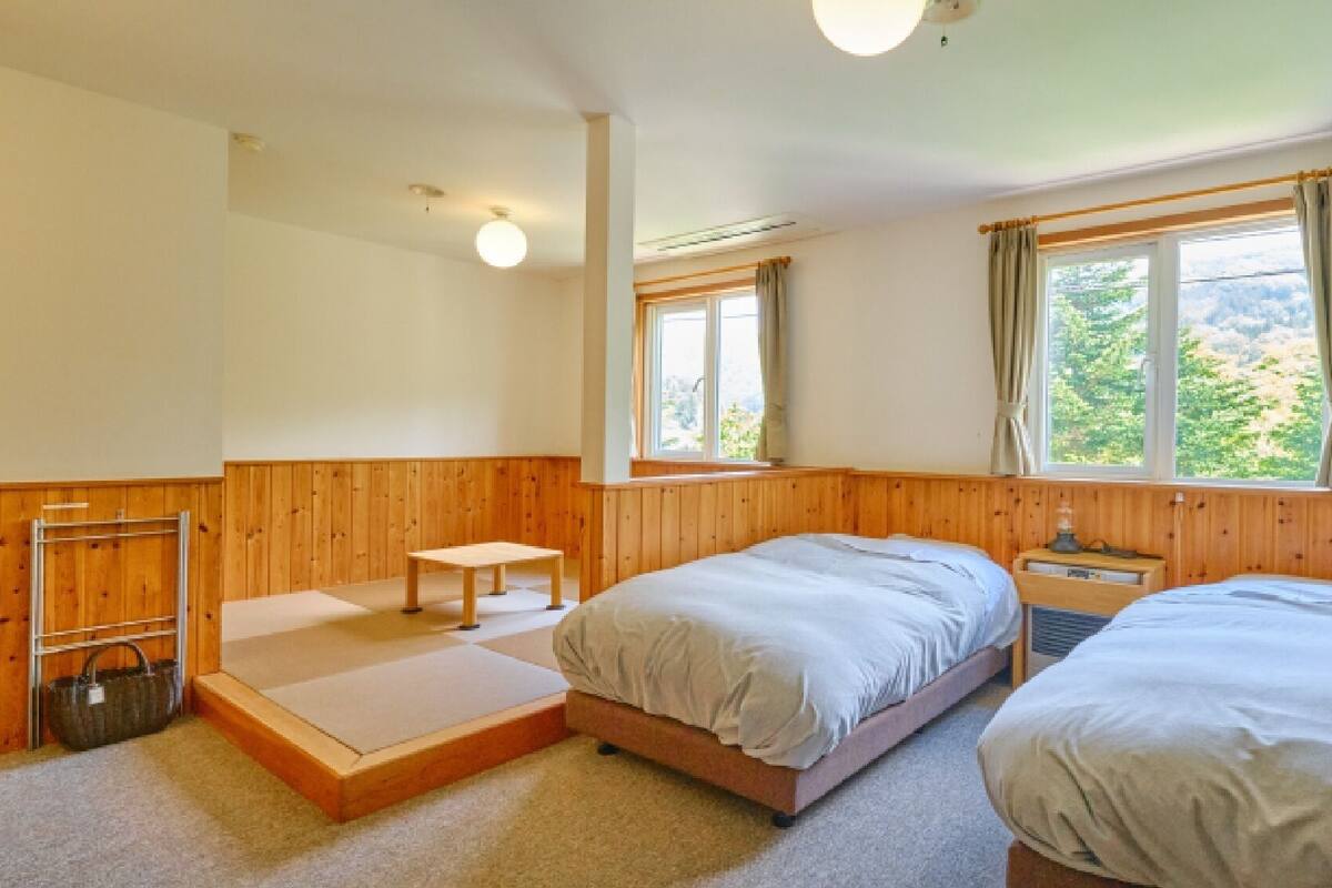 A room with two beds, a small table, a bench, and a window with curtains.