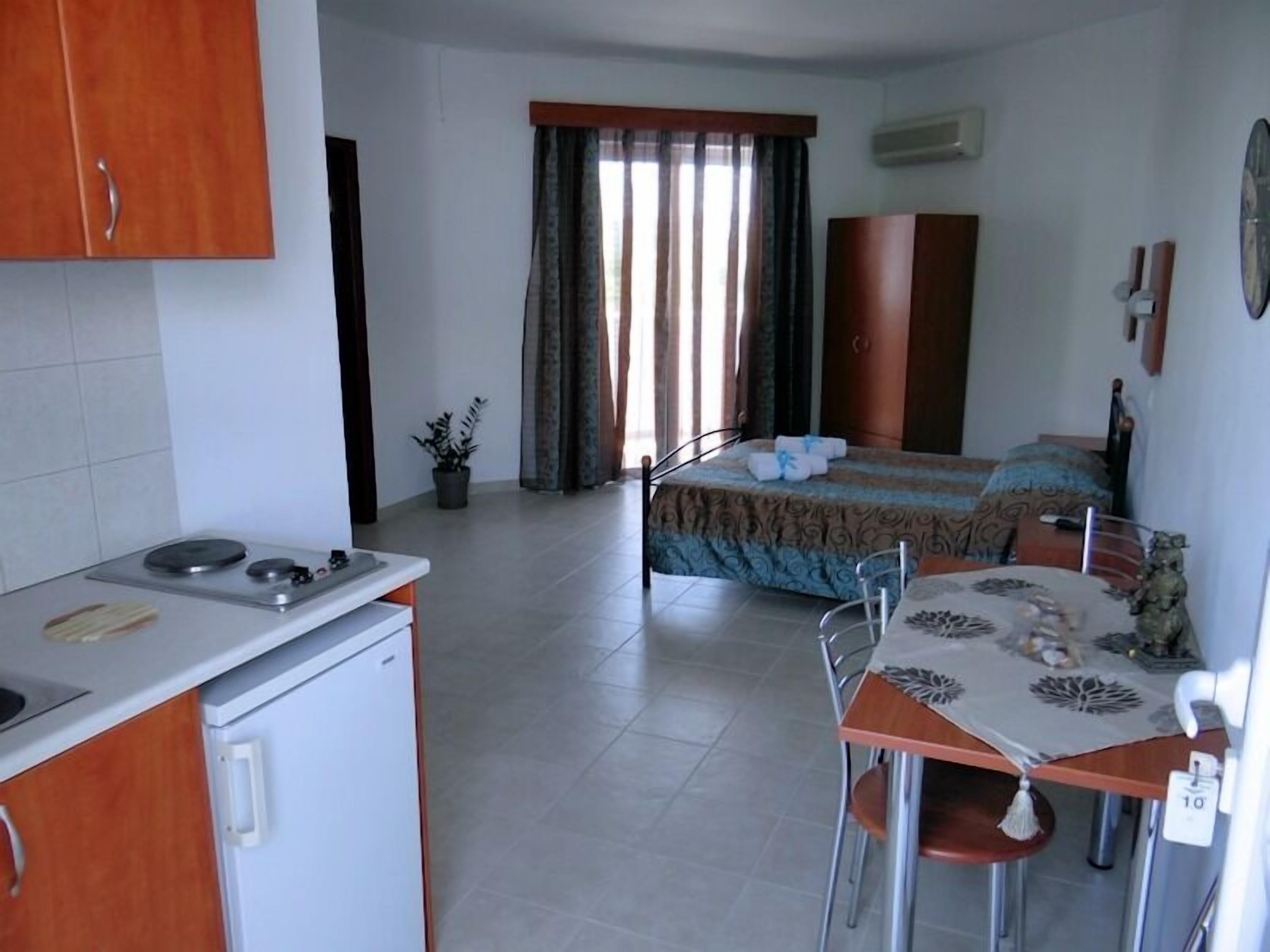 Ein kompaktes Hotelzimmer mit Küchenzeile, einem Bett mit gemustertem Überwurf, einem Esstisch mit Stühlen und einer Topfpflanze.