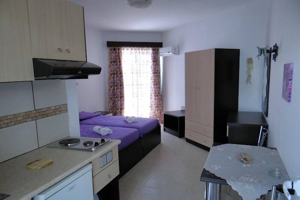 A compact hotel room with a kitchenette, a bed with purple bedding, a small table, and a wardrobe.