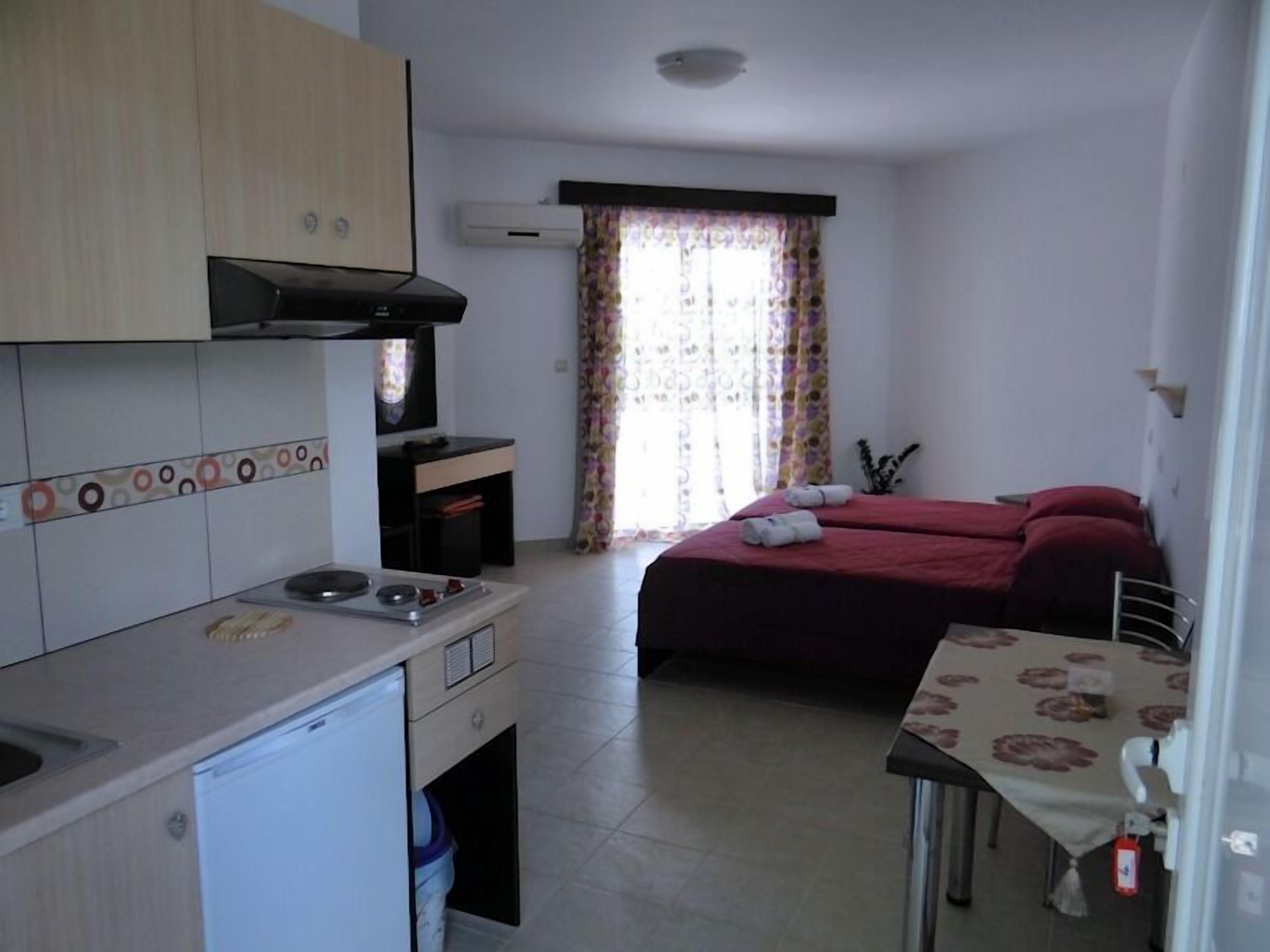 A compact hotel room with a kitchenette, a bed with red bedding, a small table, and a window with floral curtains.