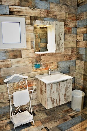 A bathroom with a rustic wooden wall, a white sink, a mirror, a hanging towel rack, and a trash bin. - Anelia Boutique Studios & Apartments (Rhodes)