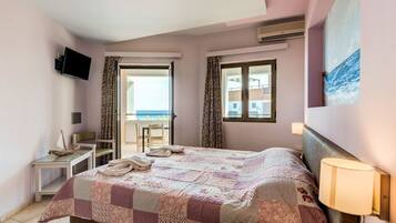 A bedroom with a bed, a TV, a desk, and a chair. There is a balcony with a view of the sea.