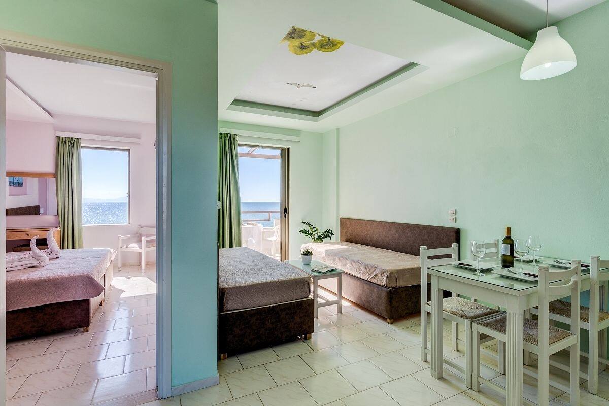 Une chambre avec deux lits, une table à manger avec des chaises, une petite table avec une bouteille et des verres, et une vue sur la mer par la fenêtre.