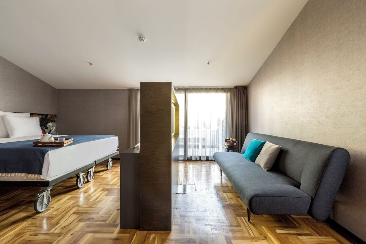 A hotel room with a bed, a sofa, a wooden floor, and a large window with curtains.