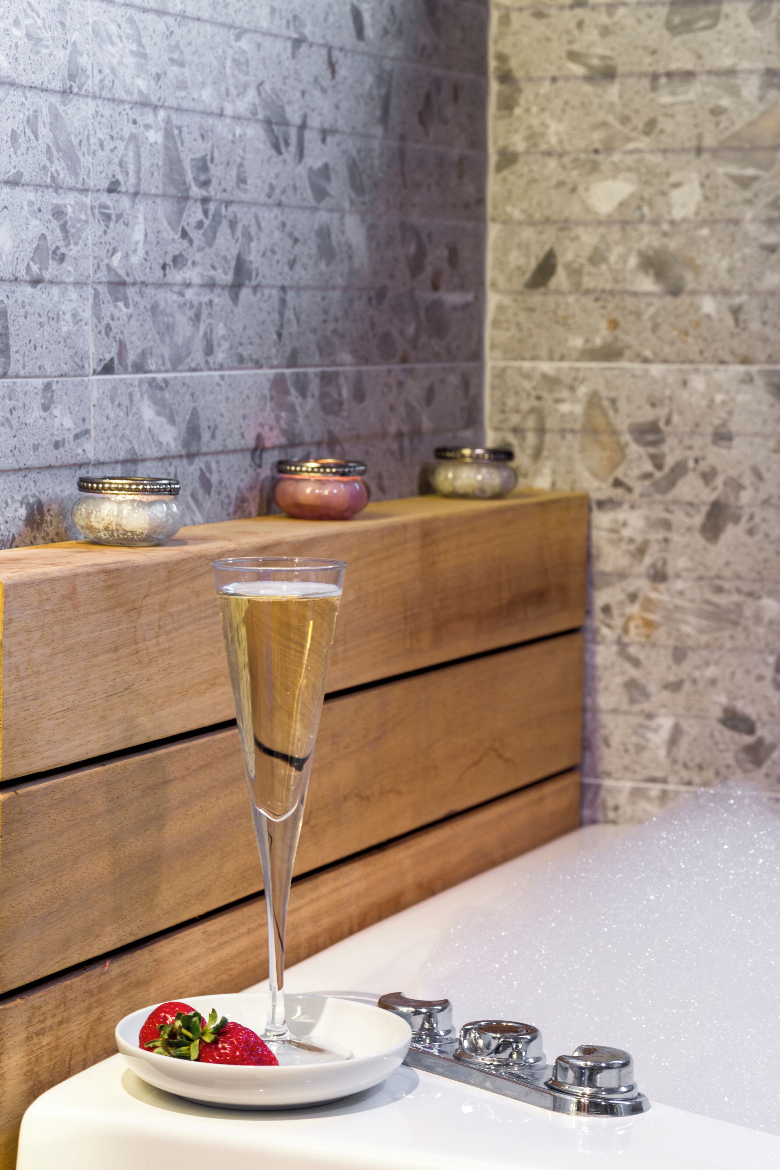 A glass of champagne on a wooden bench with a plate of strawberries, a candle, and a decorative bowl.