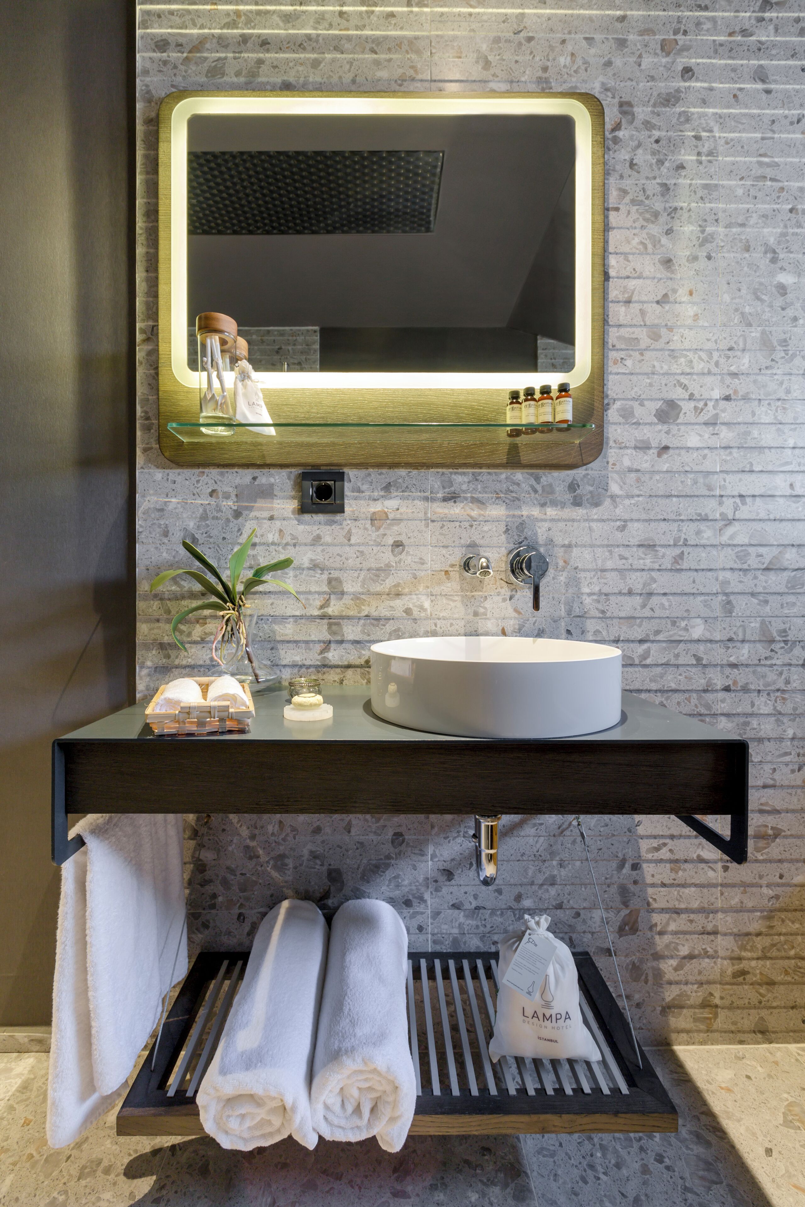 A modern bathroom with a round sink, a mirror with a light, a towel rack, and a small plant.