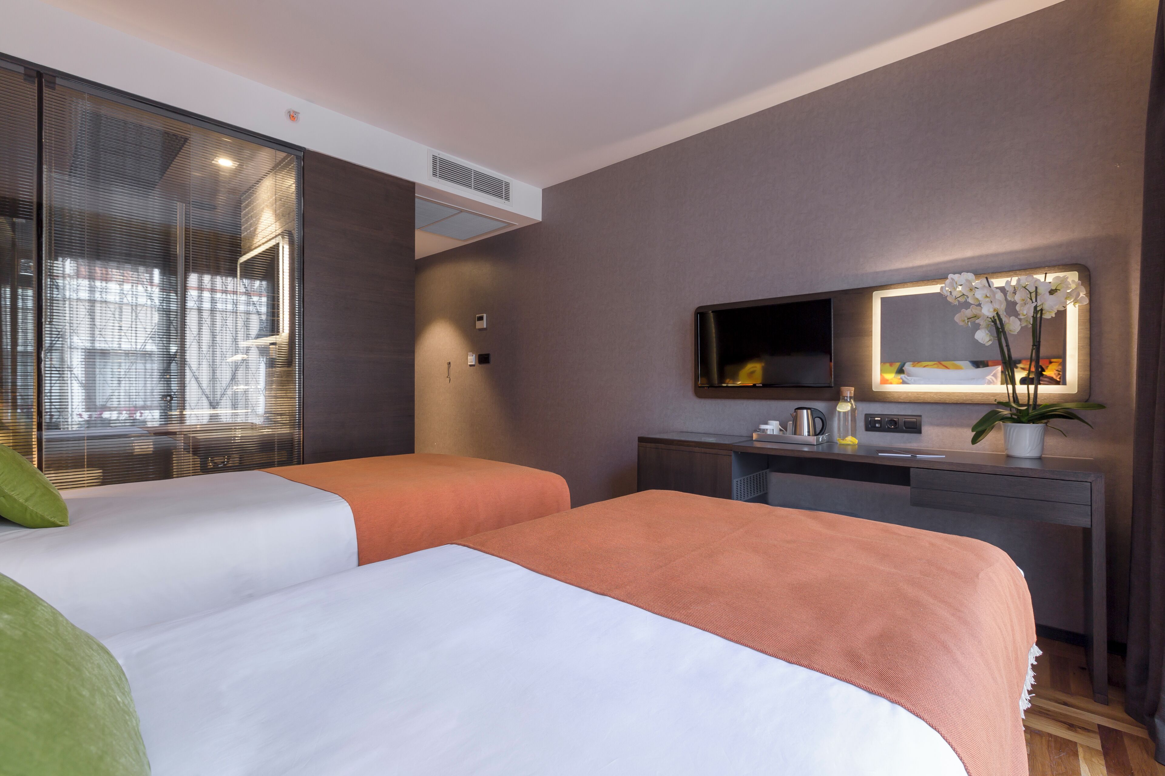A hotel room with two beds, a desk, a mirror, and a potted plant.