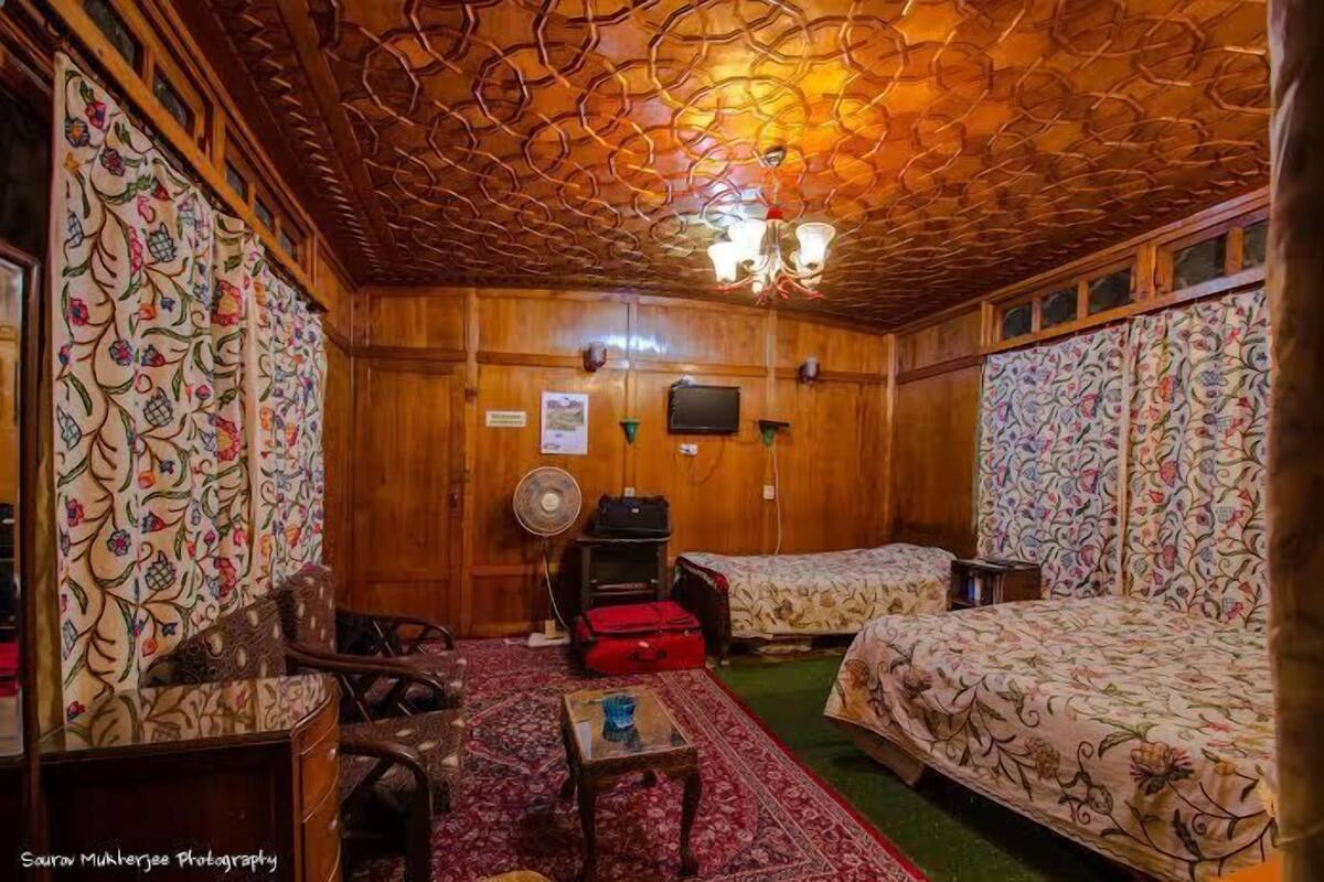 Quarto com paredes de madeira, duas camas com roupa de cama floral, uma mesa pequena, um ventilador e uma mala vermelha.