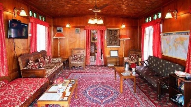 A traditional room with a wooden ceiling, a large red patterned carpet, and a television mounted on the wall. - Lily of Nageen (Srinagar)