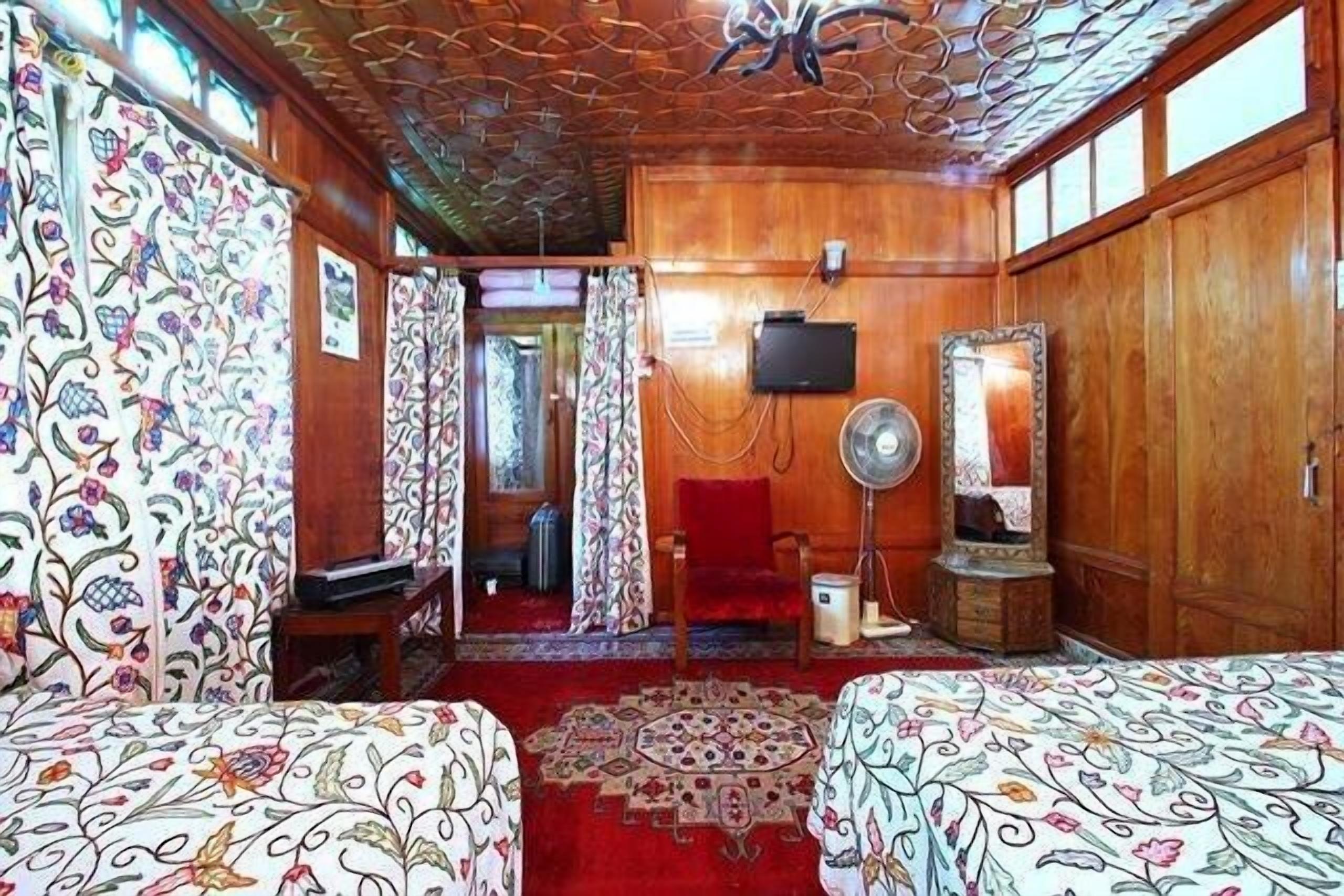 Quarto com paredes de madeira, carpete estampado, duas camas com cabeceiras combinando, ventilador de teto, espelho e televisão.