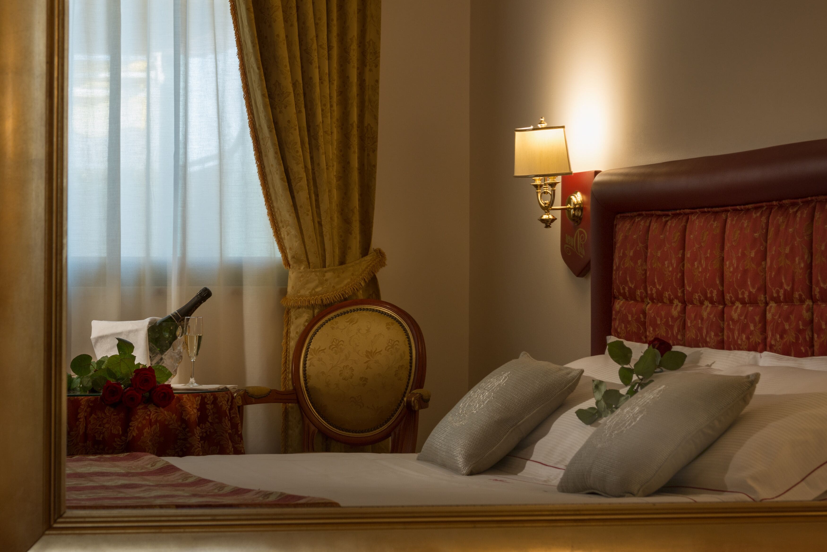 Quarto de hotel com cama, mesa de cabeceira com garrafa e copo, uma cadeira e luminária fixada na parede.