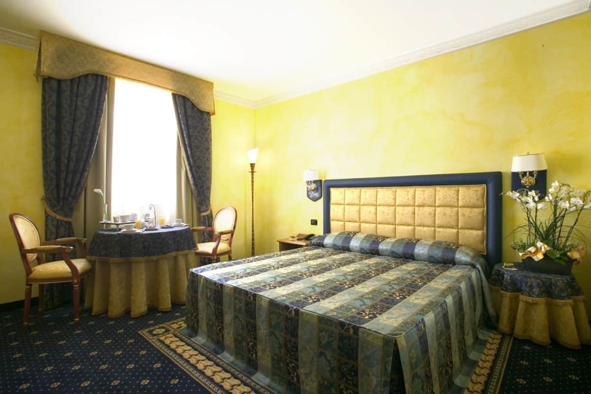 A hotel room with a bed, a round table with chairs, a lamp, and a potted plant.