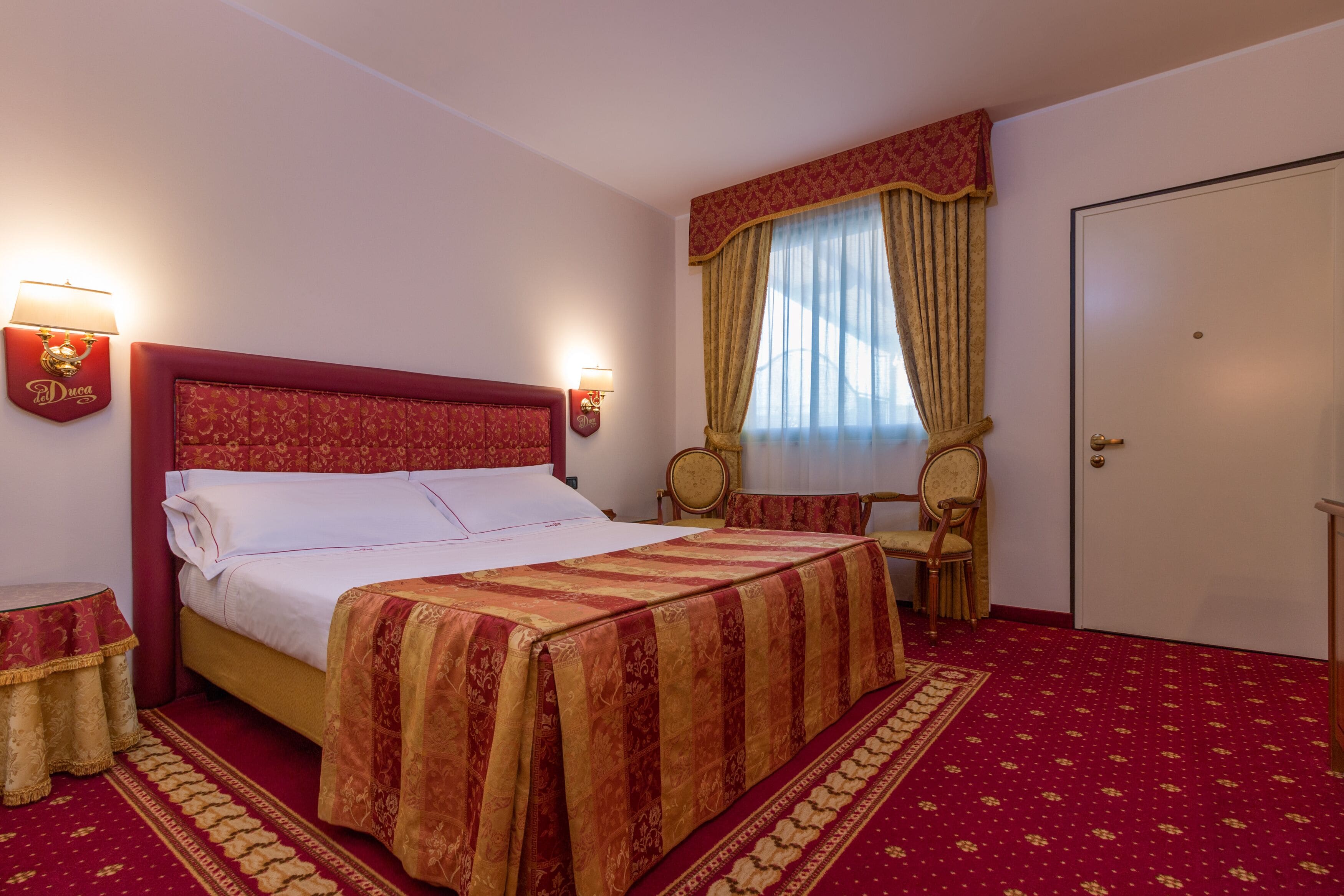 Une chambre d’hôtel avec un grand lit, deux tables de chevet, une chaise, une armoire et une fenêtre avec des rideaux.