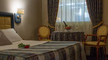 Habitación de hotel con una cama, una mesa con una botella y vasos, dos sillas y una ventana con cortinas.