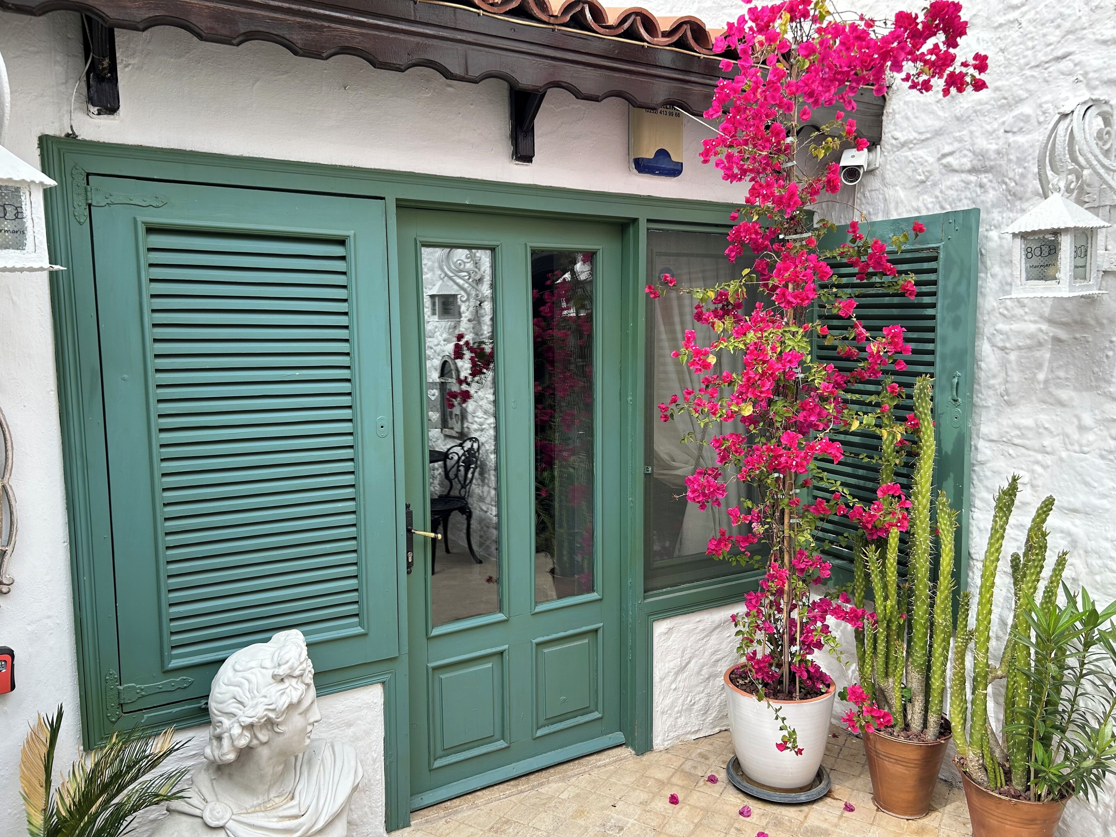 Um edifício com persianas verdes, parede branca e um vaso com flores cor-de-rosa.