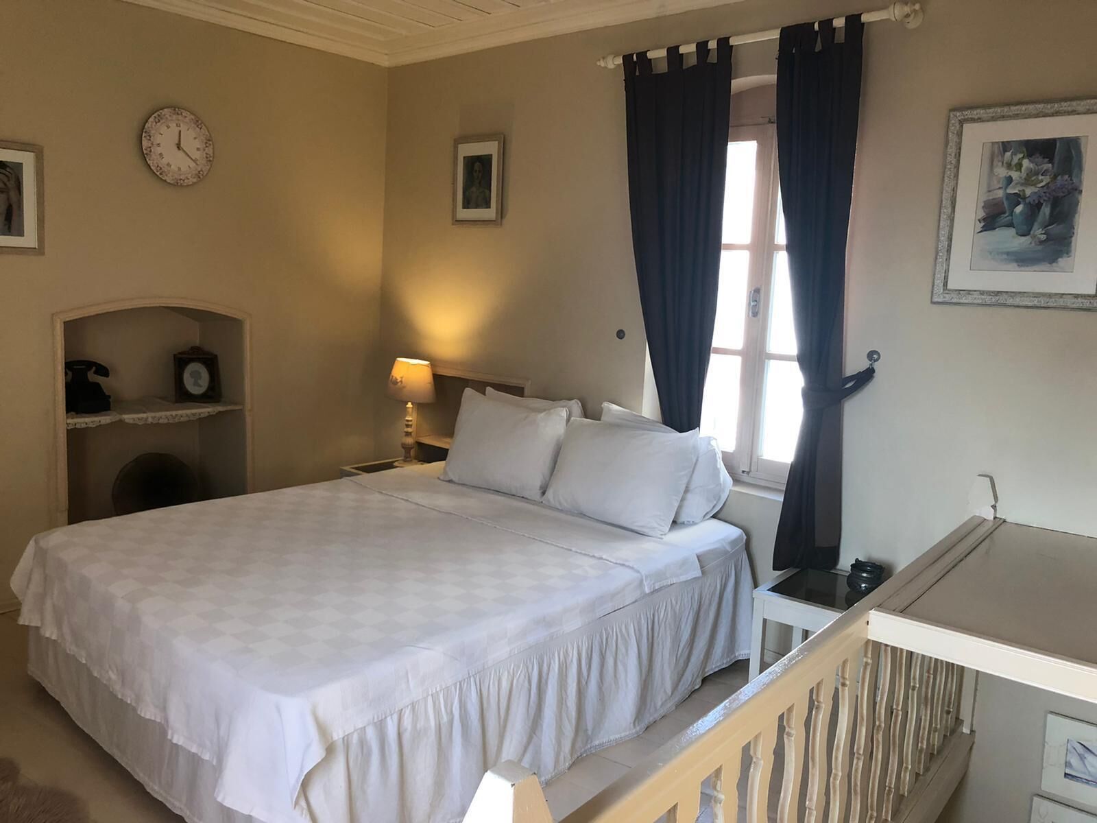 Ein Schlafzimmer mit einem Bett, einem Nachttisch, einer Lampe, einer Uhr, einem gerahmten Bild und einem Fenster mit Vorhängen.
