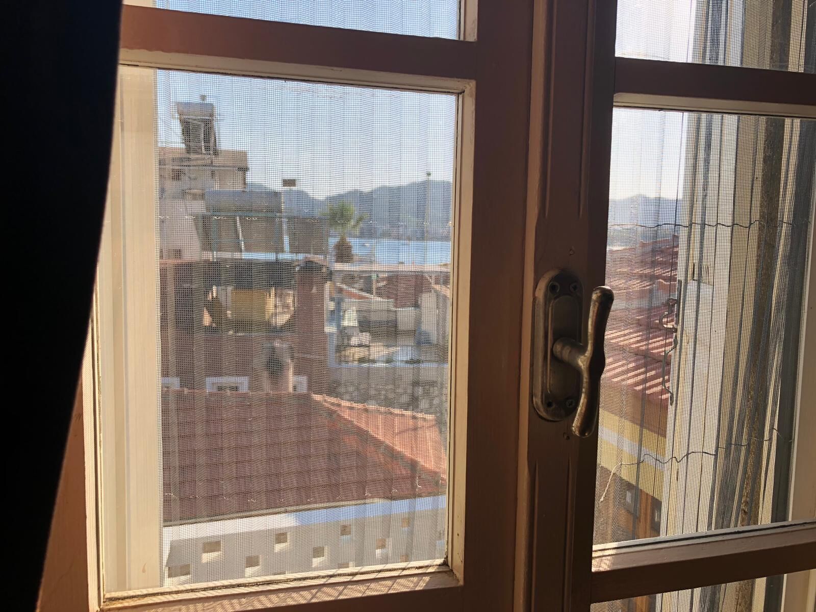 A view through a window with a mesh screen, revealing a building with a red roof and a balcony.