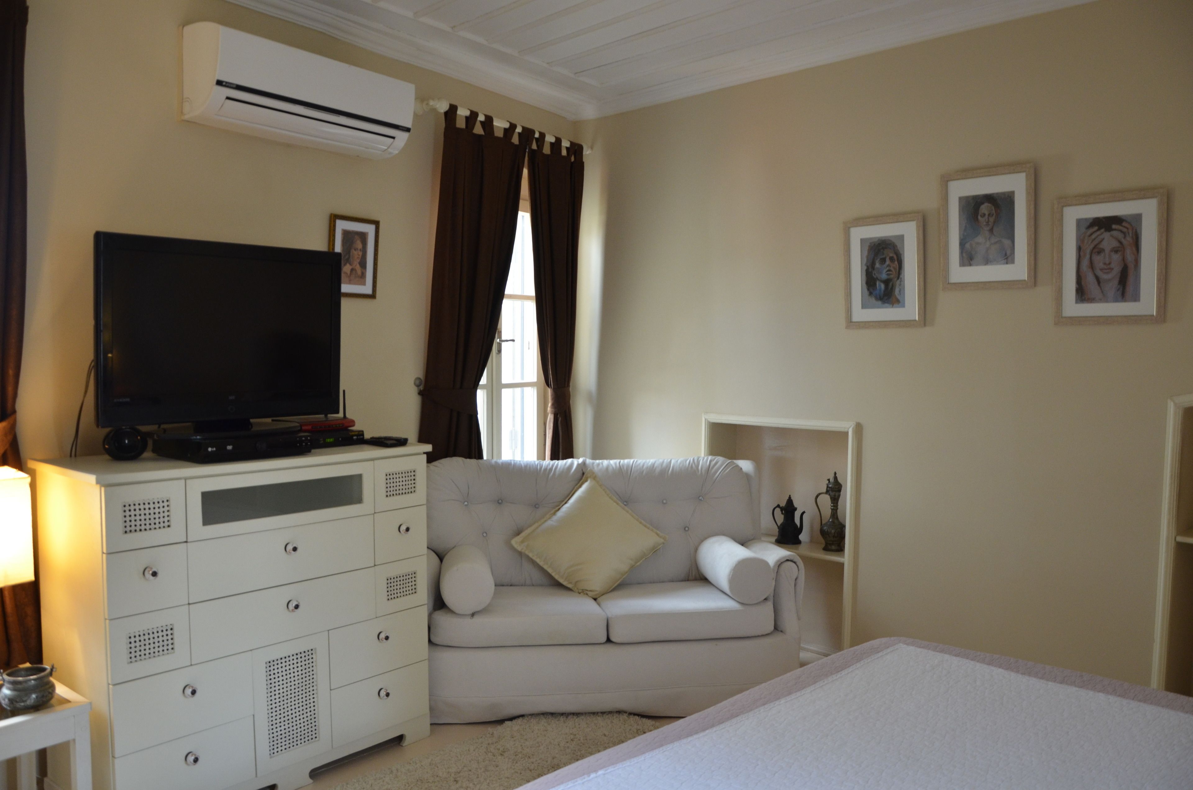 Une chambre avec un canapé blanc, une télévision, une commode et des tableaux encadrés accrochés au mur.