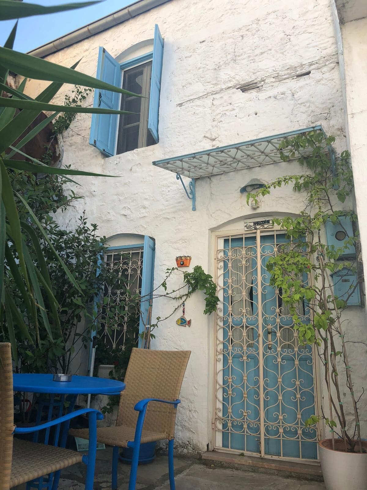 Un edificio bianco con persiane blu e un cancello in metallo. All'esterno, sedie in vimini e un tavolo.