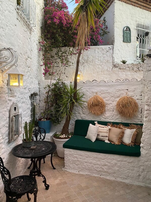 Un área de estar al aire libre acogedora, con un sofá verde, una mesa y sillas, rodeada de paredes blancas con textura y plantas en macetas.