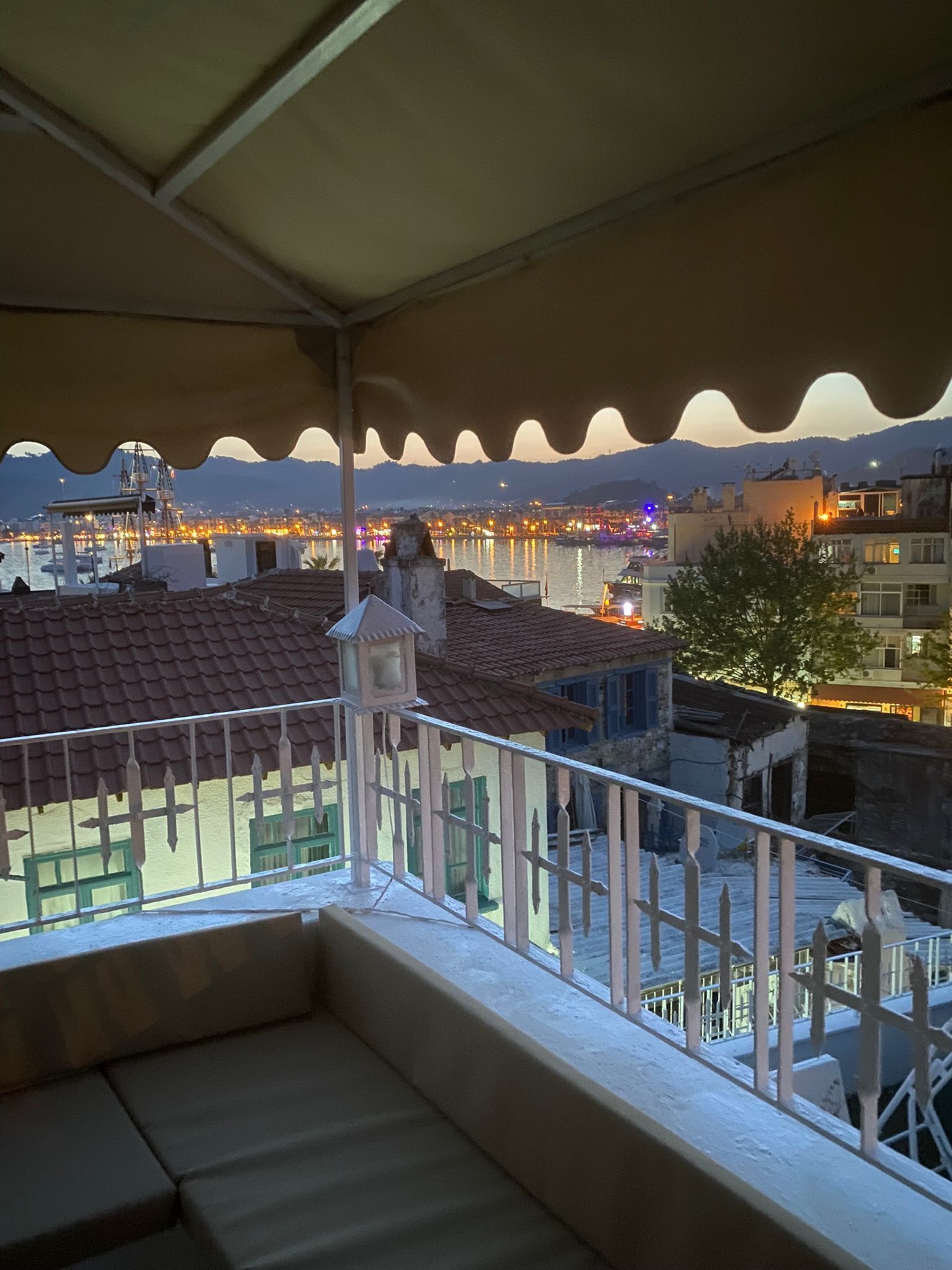 A balcony with a view of a cityscape at dusk, featuring buildings with red-tiled roofs and a body of water in the background.