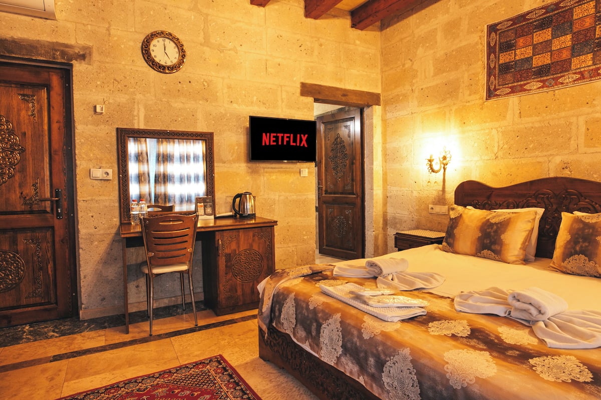 Quarto de hotel com cama de madeira, uma mesa com cadeira, uma televisão exibindo 'NETFLIX' e um relógio na parede.