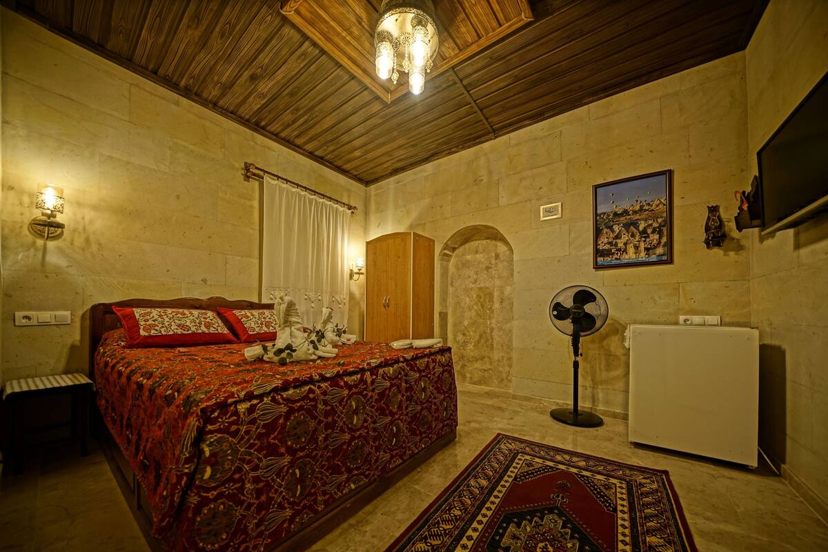 Um quarto com teto de madeira, uma cama com colcha vermelha estampada, um ventilador de chão, um radiador branco e uma TV fixada na parede.