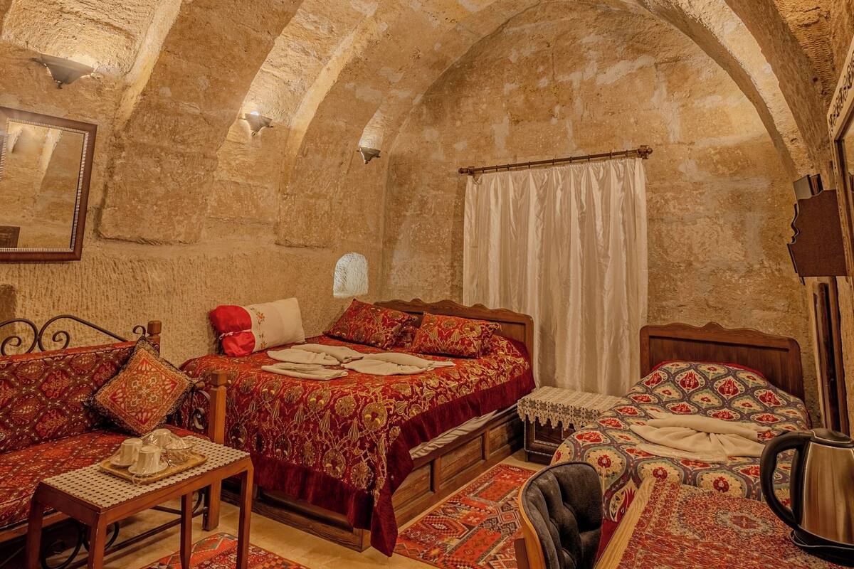 Quarto com teto abobadado, cama com colcha estampada em vermelho e branco, mesa de madeira e tapete estampado.