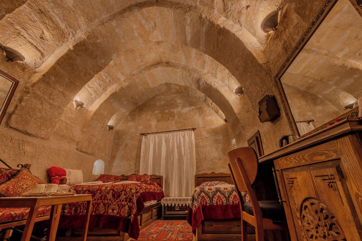 Quarto com tetos de pedra em arco, duas camas com colchas estampadas, uma mesa de madeira com bandeja e uma cadeira.