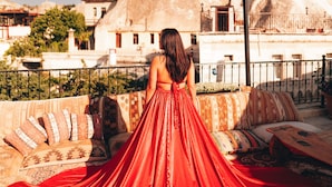 Una donna con un abito rosso seduta su un balcone che si affaccia su un edificio con due grandi strutture piramidali.
